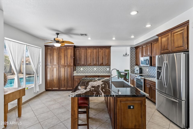 kitchen with sink, a center island with sink, light tile patterned floors, dark stone counters, and stainless steel appliances