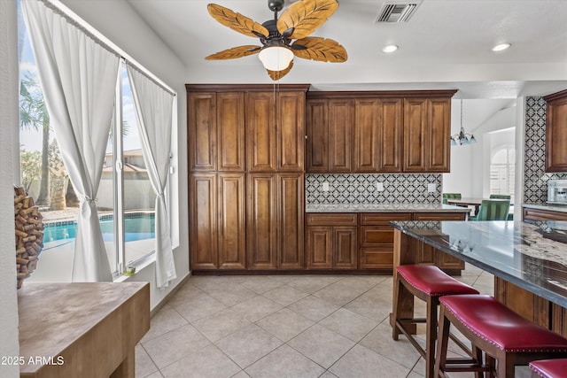 kitchen with backsplash, a kitchen bar, dark stone counters, light tile patterned floors, and ceiling fan