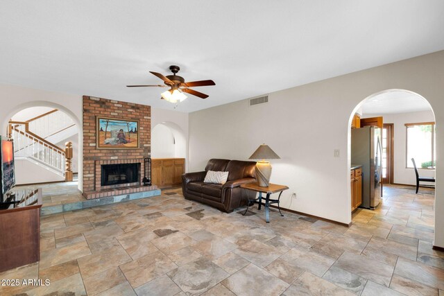 living room with stairway, a ceiling fan, baseboards, visible vents, and a brick fireplace