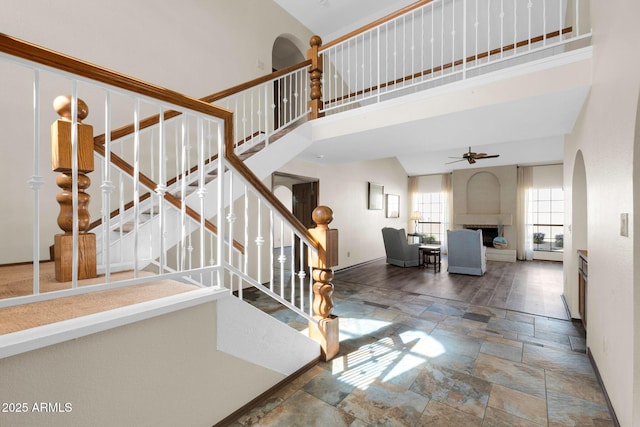 staircase featuring a fireplace with raised hearth, a ceiling fan, stone tile flooring, baseboards, and a towering ceiling