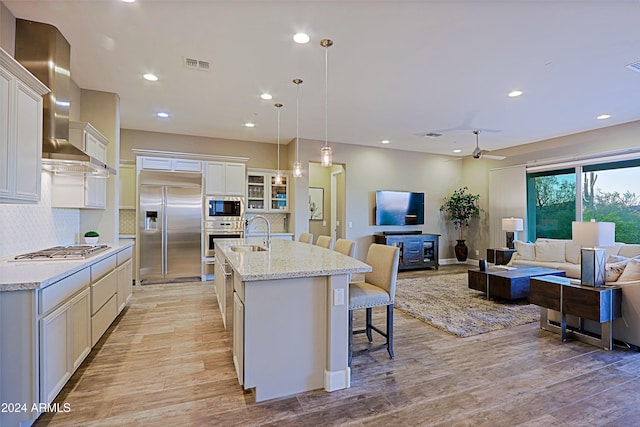 kitchen with light wood-type flooring, built in appliances, a kitchen island with sink, and wall chimney range hood