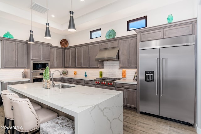 kitchen with a kitchen island with sink, built in appliances, light stone counters, sink, and a breakfast bar area