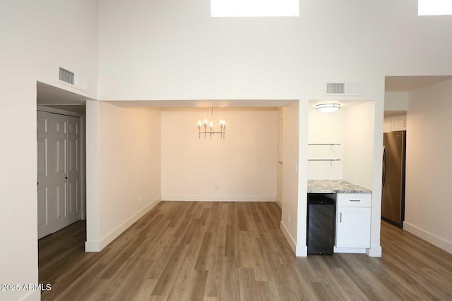 unfurnished dining area featuring beverage cooler, a chandelier, wood finished floors, baseboards, and a towering ceiling
