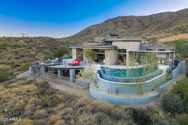 exterior space featuring a patio, a mountain view, fence, stone siding, and an infinity pool