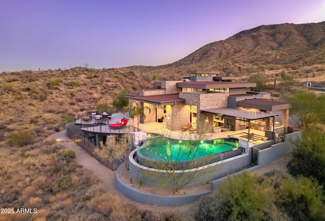 back of property at dusk featuring stone siding, an infinity pool, a mountain view, and a patio