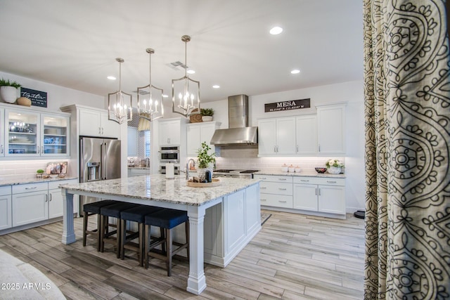 kitchen featuring a center island with sink, stainless steel appliances, white cabinets, wall chimney range hood, and glass insert cabinets