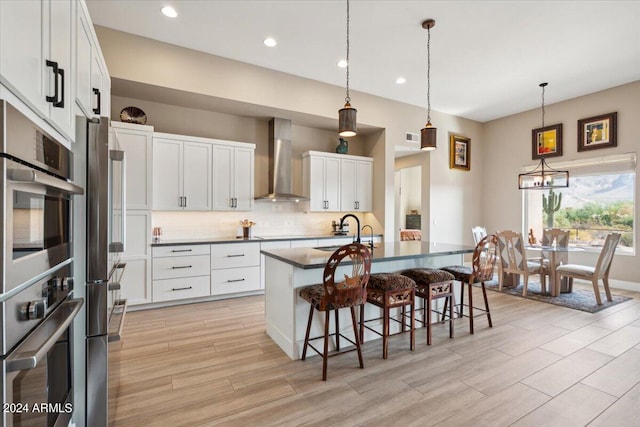 kitchen featuring white cabinets, a center island with sink, double oven, and wall chimney range hood