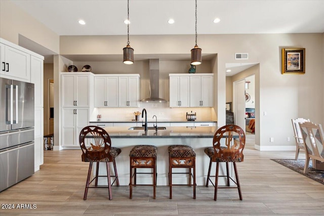 kitchen with sink, white cabinets, wall chimney range hood, stainless steel refrigerator, and a center island with sink