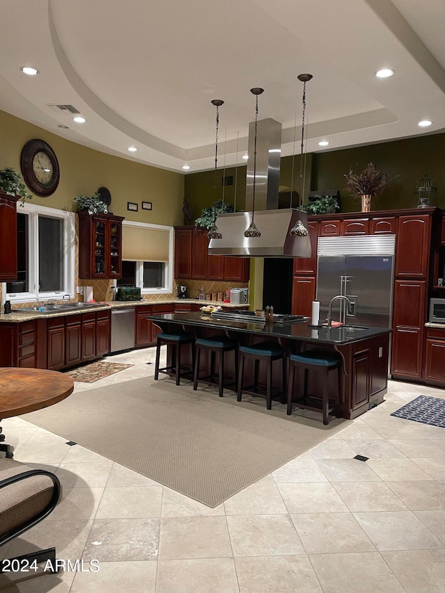 kitchen featuring a kitchen island with sink, island range hood, stainless steel appliances, a raised ceiling, and light colored carpet