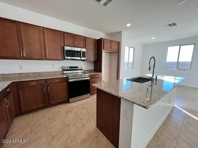 kitchen with appliances with stainless steel finishes, light stone counters, a kitchen island with sink, sink, and light tile patterned floors