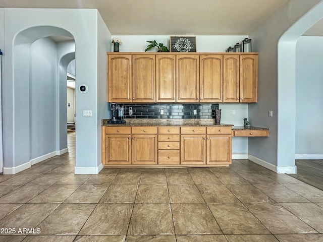 kitchen featuring arched walkways, tile patterned flooring, baseboards, dark stone counters, and tasteful backsplash