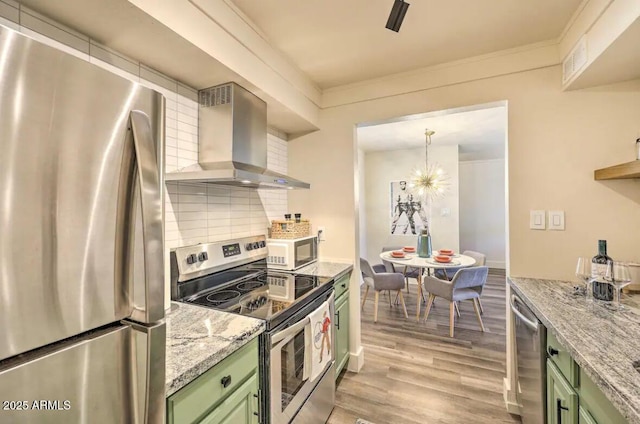 kitchen with green cabinetry, light wood-style flooring, stainless steel appliances, wall chimney range hood, and backsplash