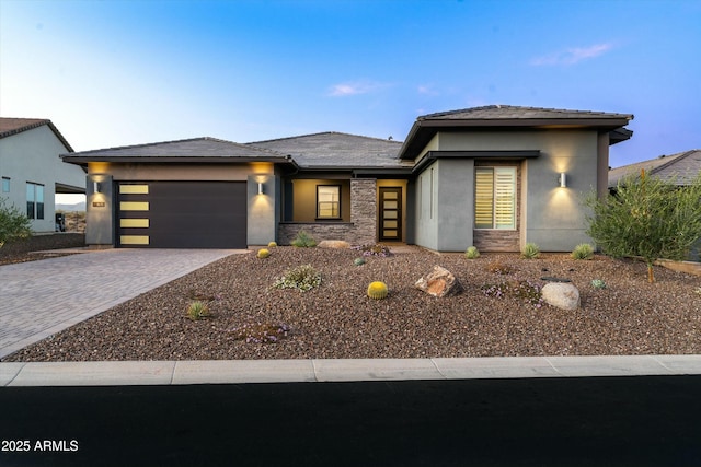 prairie-style house with stone siding, decorative driveway, an attached garage, and stucco siding