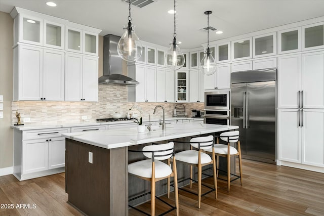 kitchen with wall chimney exhaust hood, glass insert cabinets, built in appliances, a kitchen island with sink, and white cabinetry