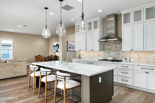 kitchen with visible vents, glass insert cabinets, a kitchen island with sink, white cabinetry, and wall chimney exhaust hood