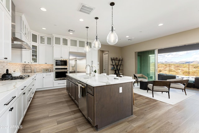 kitchen featuring a center island with sink, visible vents, glass insert cabinets, white cabinetry, and built in appliances