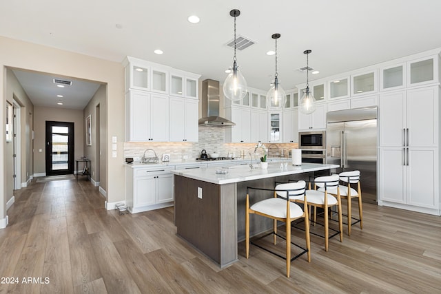 kitchen with built in appliances, wall chimney range hood, glass insert cabinets, and a center island with sink