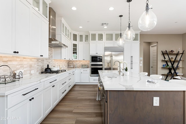 kitchen featuring white cabinets, wall chimney exhaust hood, built in appliances, hanging light fixtures, and a kitchen island with sink