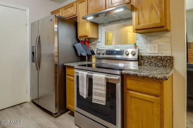 kitchen featuring stainless steel appliances, dark stone countertops, under cabinet range hood, light wood finished floors, and decorative backsplash