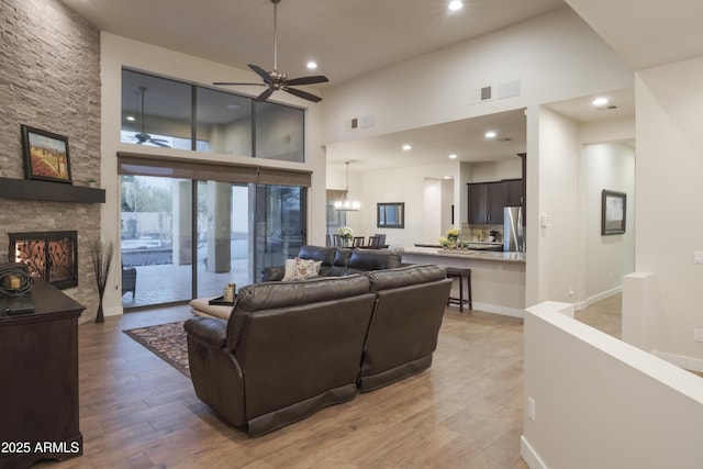 living room featuring ceiling fan with notable chandelier, light hardwood / wood-style floors, a stone fireplace, and a towering ceiling