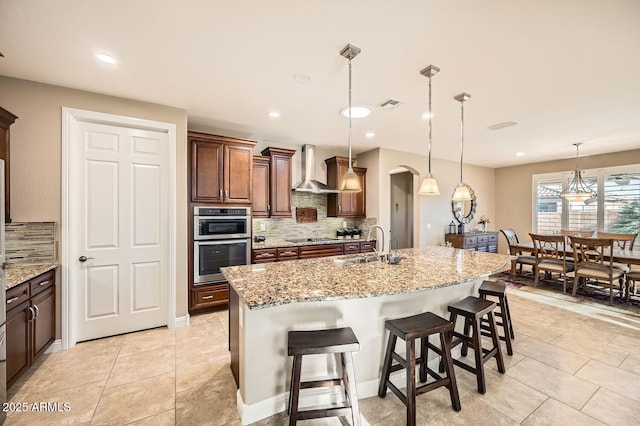 kitchen featuring wall chimney exhaust hood, a center island with sink, arched walkways, and decorative light fixtures