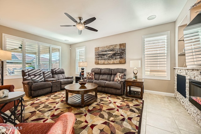 living area featuring light tile patterned floors, a glass covered fireplace, a ceiling fan, and baseboards