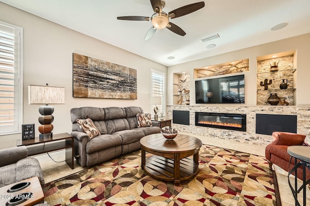 living room with built in shelves, a glass covered fireplace, visible vents, and ceiling fan