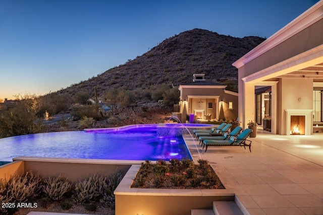 pool at dusk with a mountain view, pool water feature, exterior fireplace, and a patio area