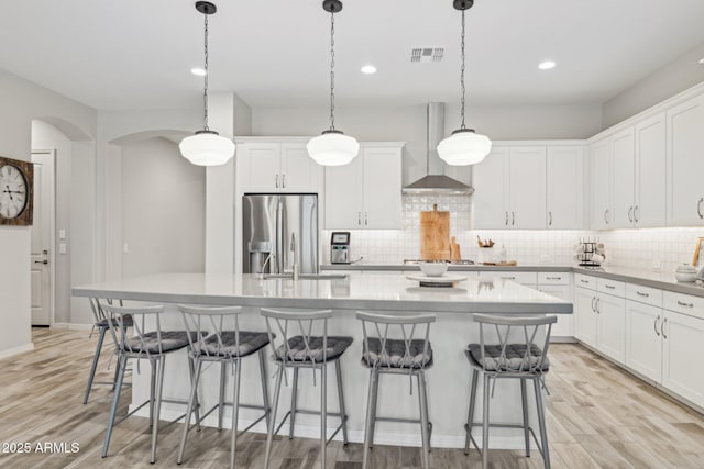 kitchen featuring a center island with sink, arched walkways, light wood-style flooring, appliances with stainless steel finishes, and wall chimney range hood