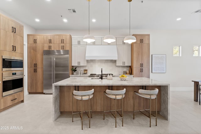 kitchen with light stone countertops, custom exhaust hood, decorative light fixtures, stainless steel appliances, and an island with sink