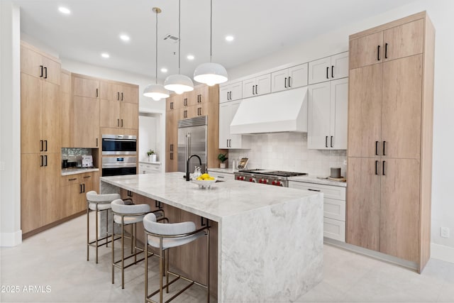 kitchen with light brown cabinets, tasteful backsplash, premium range hood, light stone countertops, and a kitchen island with sink