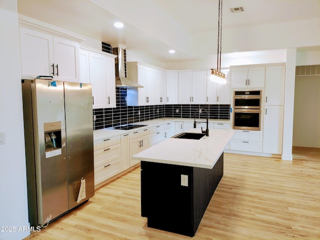 kitchen featuring decorative light fixtures, an island with sink, sink, stainless steel appliances, and wall chimney range hood