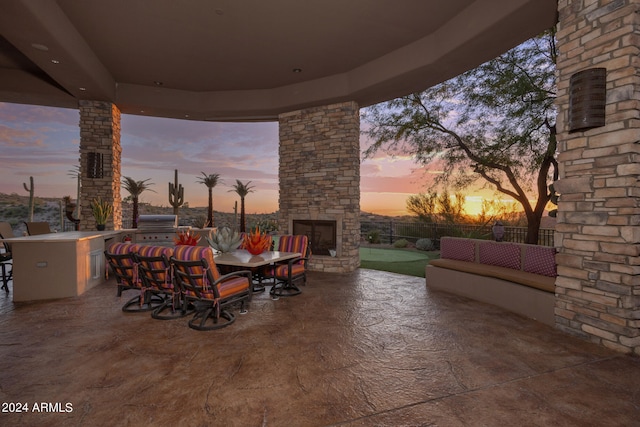 patio terrace at dusk with exterior kitchen, grilling area, and an outdoor stone fireplace