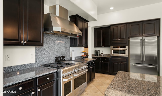 kitchen with high end appliances, light tile patterned floors, wall chimney exhaust hood, and dark stone counters