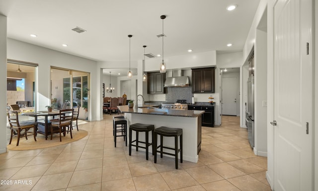 kitchen featuring pendant lighting, an island with sink, wall chimney exhaust hood, light tile patterned floors, and dark brown cabinets