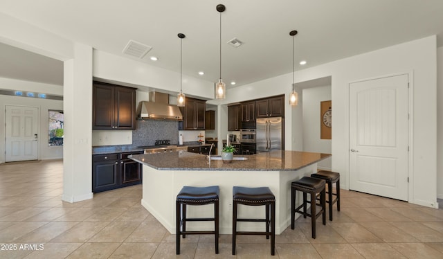 kitchen featuring appliances with stainless steel finishes, wall chimney exhaust hood, dark stone counters, an island with sink, and dark brown cabinets