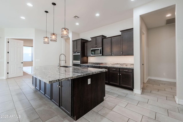 kitchen with a kitchen island with sink, light stone countertops, pendant lighting, dark brown cabinetry, and sink