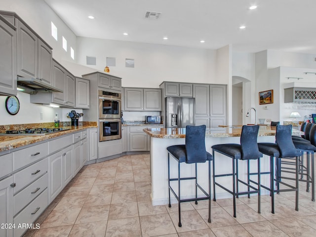 kitchen with gray cabinets, stainless steel appliances, a center island with sink, light stone countertops, and a high ceiling