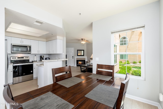 dining area featuring a ceiling fan, plenty of natural light, a lit fireplace, and baseboards