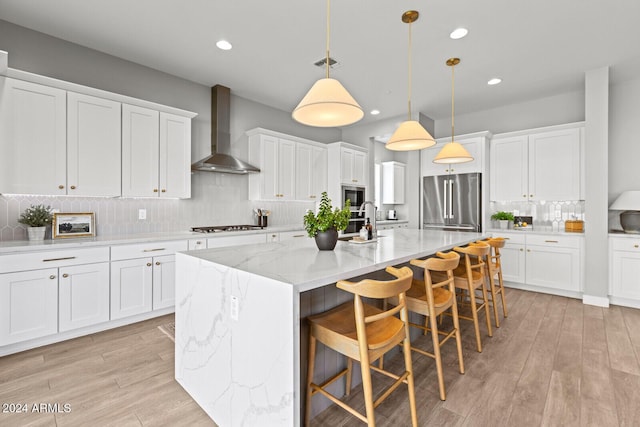 kitchen with stainless steel appliances, a kitchen island with sink, wall chimney range hood, pendant lighting, and white cabinets