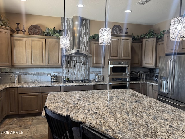 kitchen featuring light stone countertops, hanging light fixtures, appliances with stainless steel finishes, wall chimney exhaust hood, and tasteful backsplash