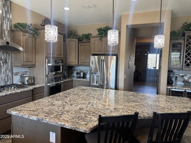 kitchen featuring wall chimney range hood, appliances with stainless steel finishes, a breakfast bar area, backsplash, and visible vents