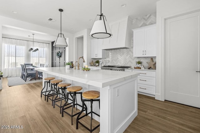 kitchen featuring custom exhaust hood, a breakfast bar area, a kitchen island with sink, pendant lighting, and white cabinets