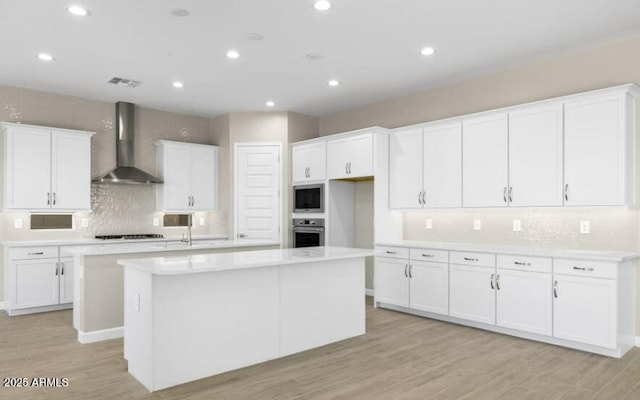 kitchen featuring white cabinetry, a center island, stainless steel appliances, light hardwood / wood-style floors, and wall chimney range hood