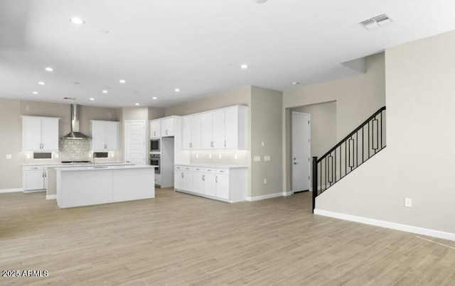 kitchen featuring wall chimney exhaust hood, tasteful backsplash, light hardwood / wood-style flooring, an island with sink, and white cabinets