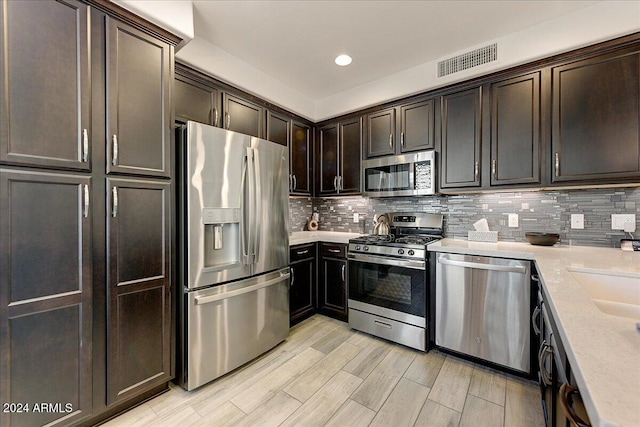 kitchen featuring dark brown cabinetry, light stone countertops, stainless steel appliances, decorative backsplash, and light wood-type flooring