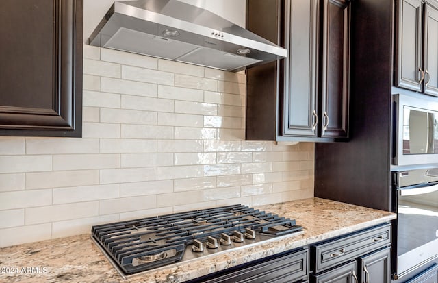 kitchen featuring wall chimney exhaust hood, backsplash, dark brown cabinetry, appliances with stainless steel finishes, and light stone counters