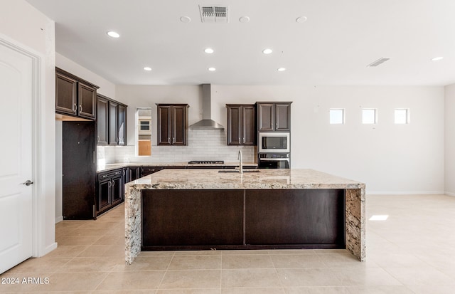 kitchen with wall chimney range hood, light stone countertops, stainless steel appliances, and an island with sink