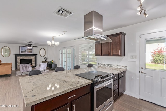 kitchen featuring stainless steel range with electric cooktop, ceiling fan with notable chandelier, a stone fireplace, island range hood, and kitchen peninsula