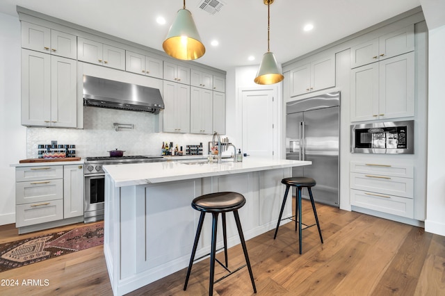 kitchen featuring backsplash, built in appliances, a center island with sink, and wood-type flooring